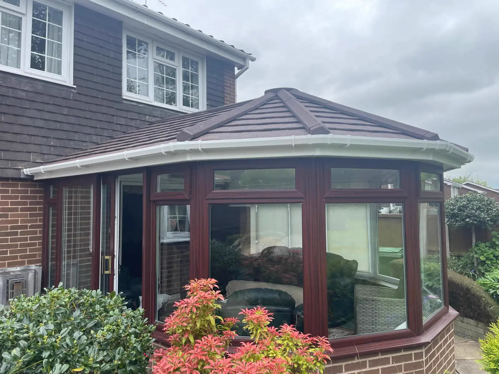 Brown-framed conservatory with a tiled roof beside flowering garden plants